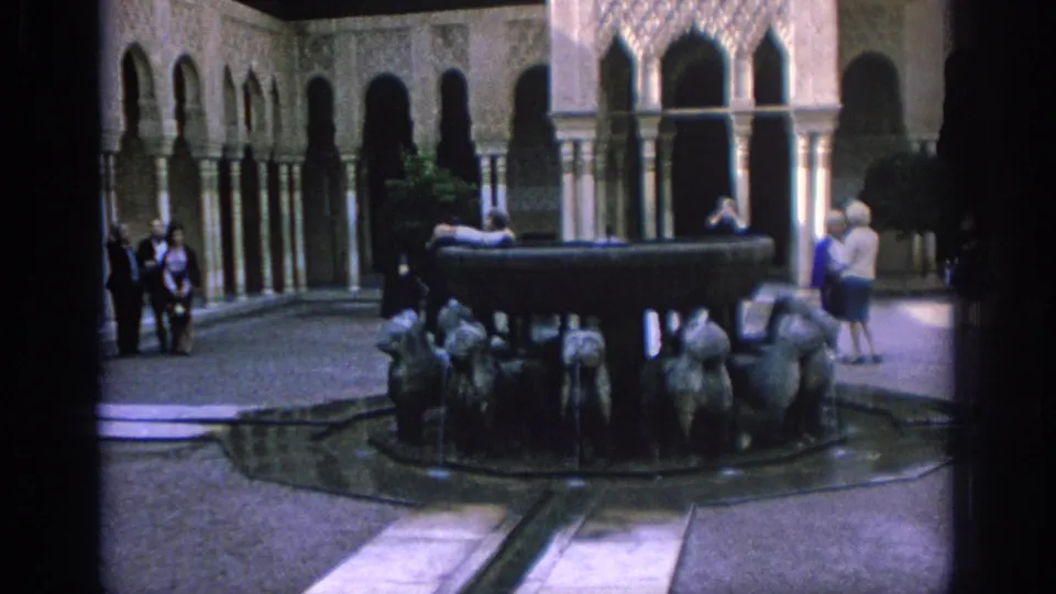 1966 Spain frame showing a courtyard fountain with sculpted animal figures, visitors nearby, and ornate arched colonnades in the background.