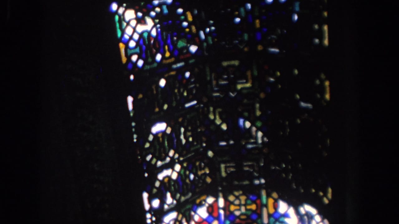 1966 Spain home-movie frame showing stained glass glowing in a dark historic interior, with geometric patterns and deep blues, reds, and greens.