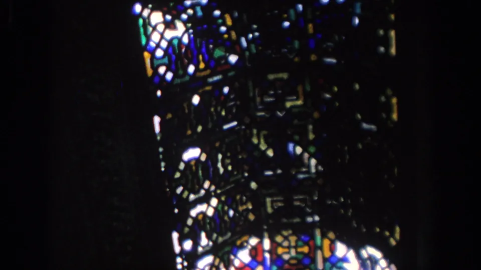 1966 Spain home-movie frame showing stained glass glowing in a dark historic interior, with geometric patterns and deep blues, reds, and greens.