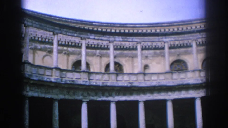 1966 Spain frame showing a curved historic facade with a colonnade, arched openings, balcony details, and soft daylight exposure.