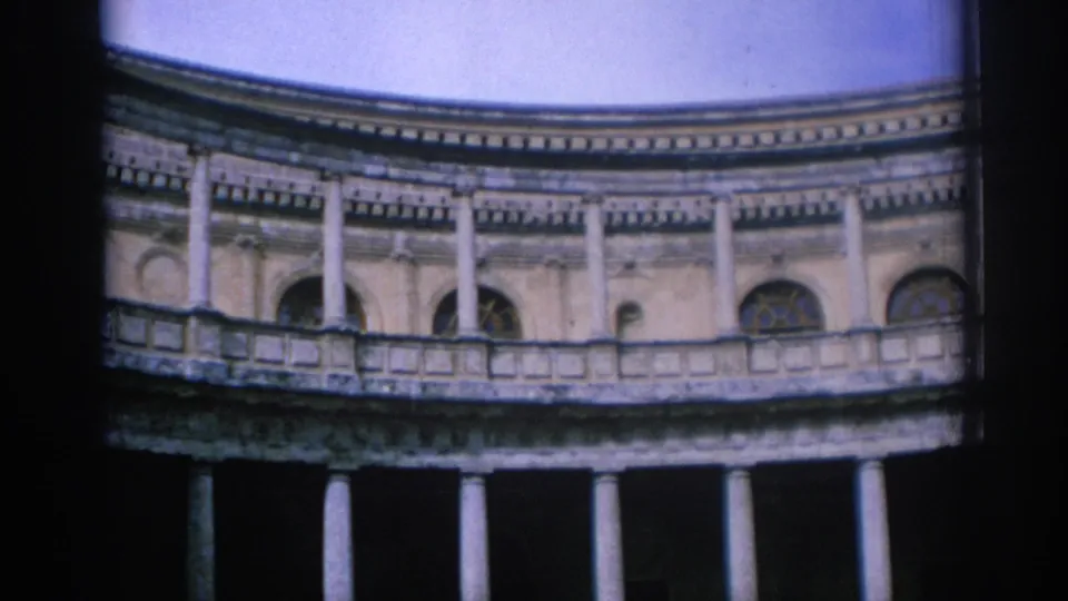 1966 Spain 8mm frame showing a low-angle view of a curved stone building with columns, balcony rail, arched windows, and blue sky.