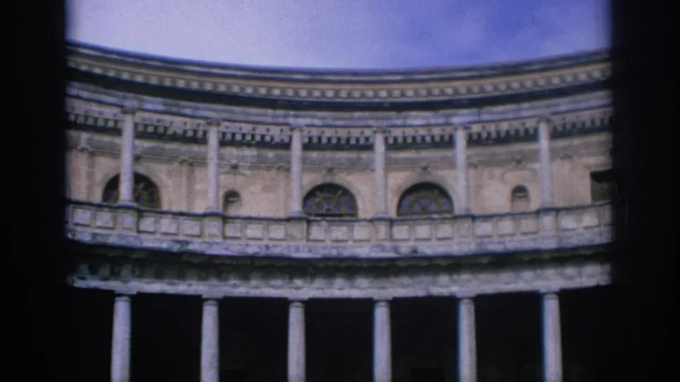 1966 Spain 8mm frame showing more of the circular facade, with upper arched windows, a stone balcony, lower columns, and blue sky.