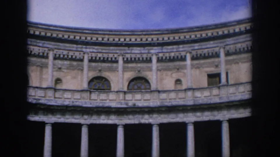 1966 Spain 8mm frame of a low-angle architectural view with columns, balcony rail, arched windows, worn stone texture, and sky.