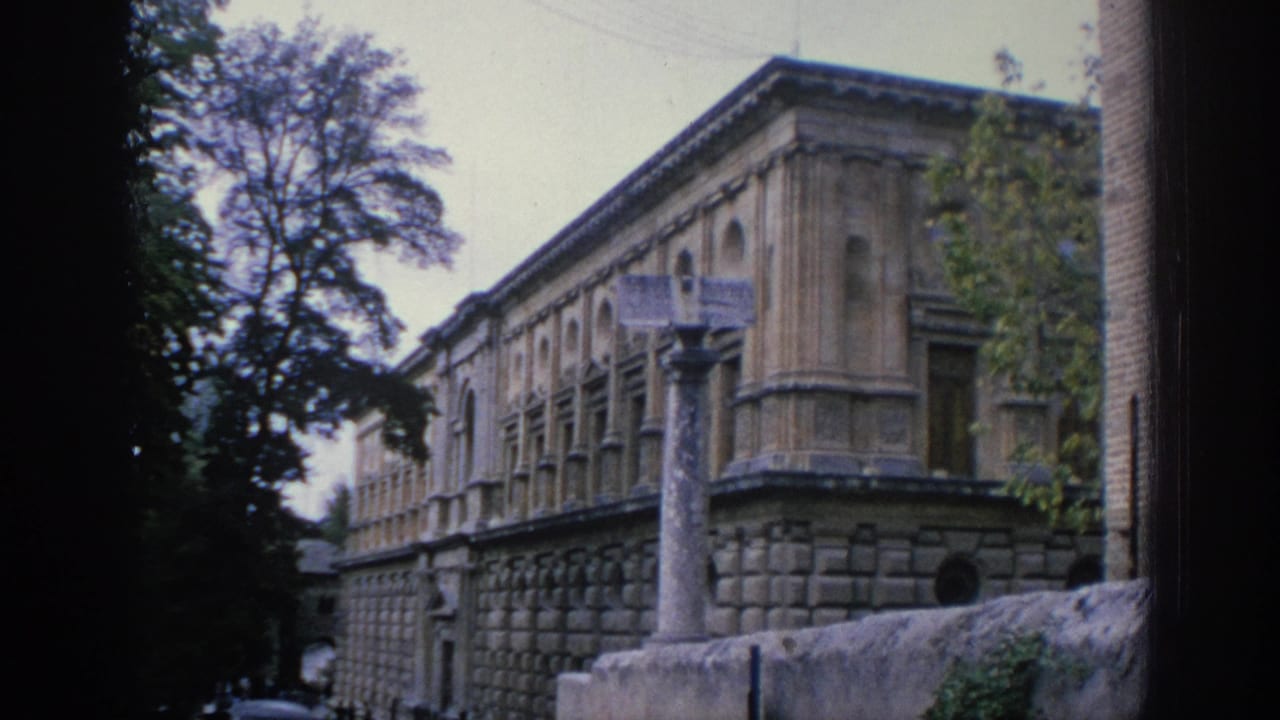 1966 Spain travel footage frame showing an ornate stone building beside trees, with a tall column and low wall in the foreground.