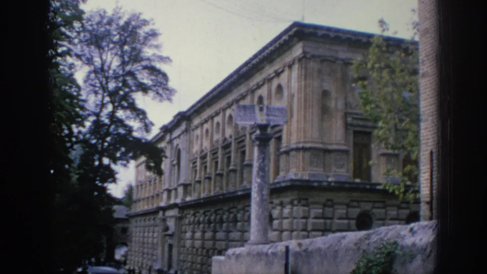 1966 Spain travel footage frame showing an ornate stone building beside trees, with a tall column and low wall in the foreground.