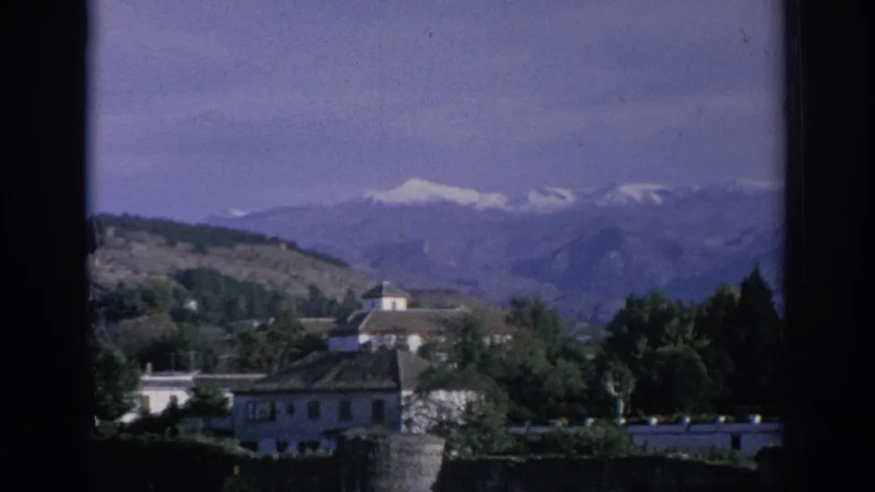 1966 Spain travel frame showing village buildings and trees in the foreground with a distant mountain range and snow-capped peaks beyond.