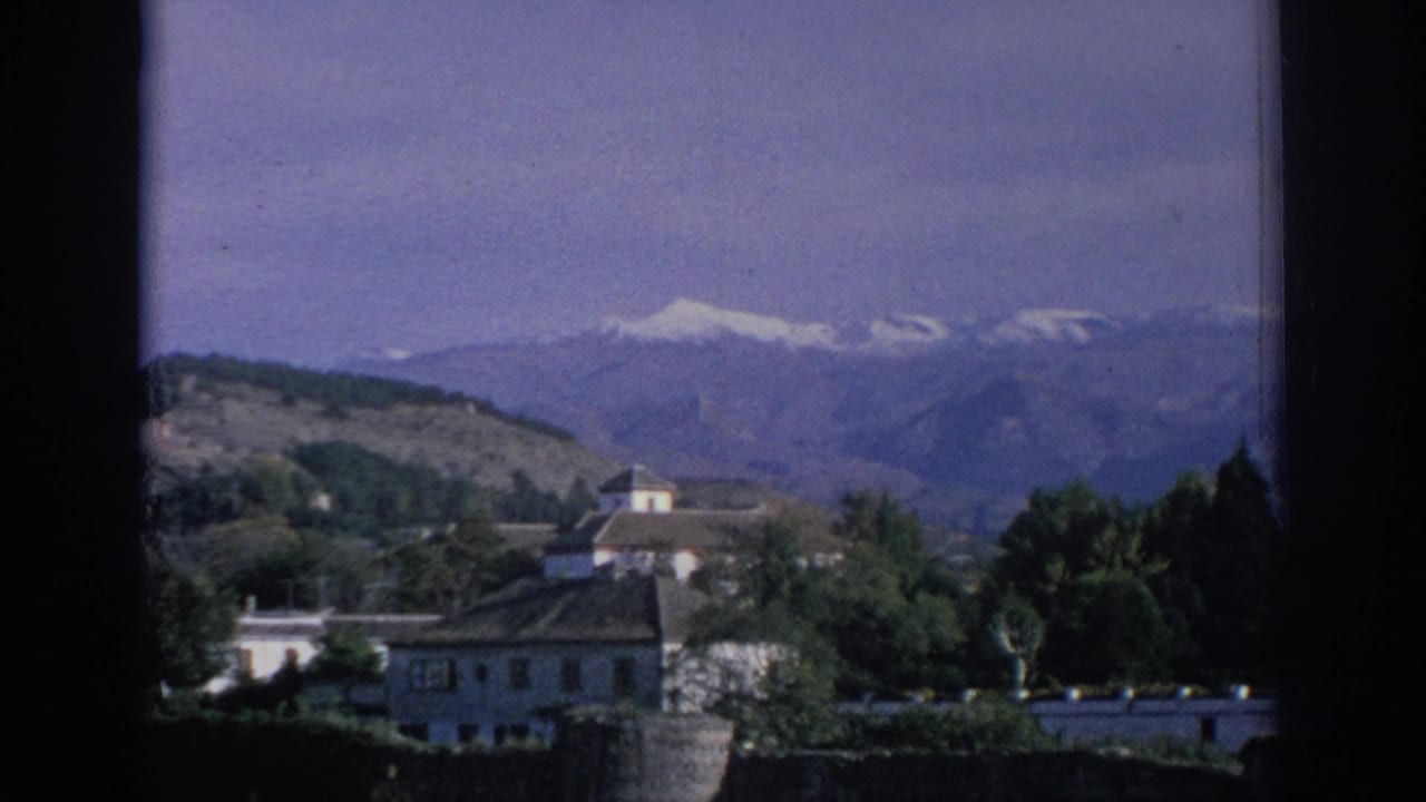 1966 Spain 8mm view of white buildings, greenery, a stone wall, and hazy snow-capped mountains beyond.