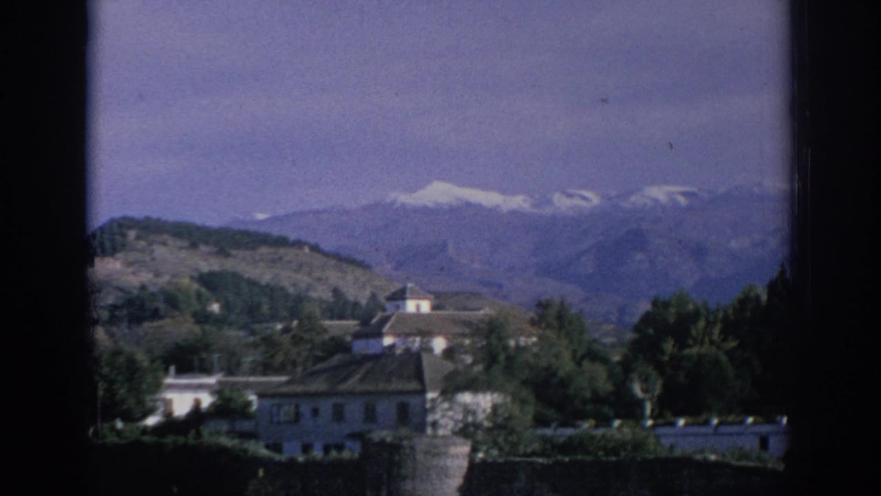 Static 1966 Spain home-movie frame showing white buildings, tree cover, and snow-capped mountains in the distance.
