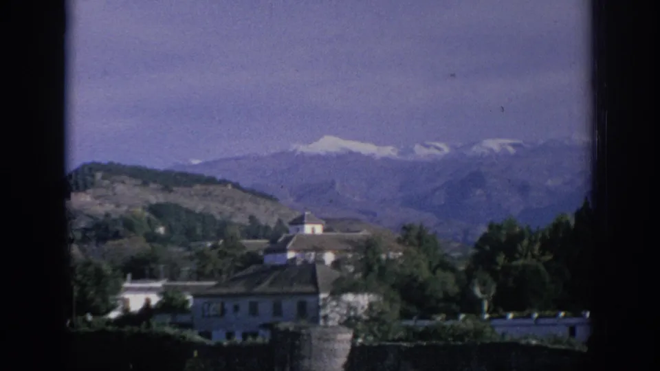 Static 1966 Spain home-movie frame showing white buildings, tree cover, and snow-capped mountains in the distance.