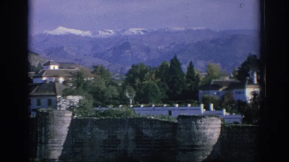 1966 Spain 8mm frame with a stone wall, white exterior buildings, dense trees, and a hazy mountain backdrop.