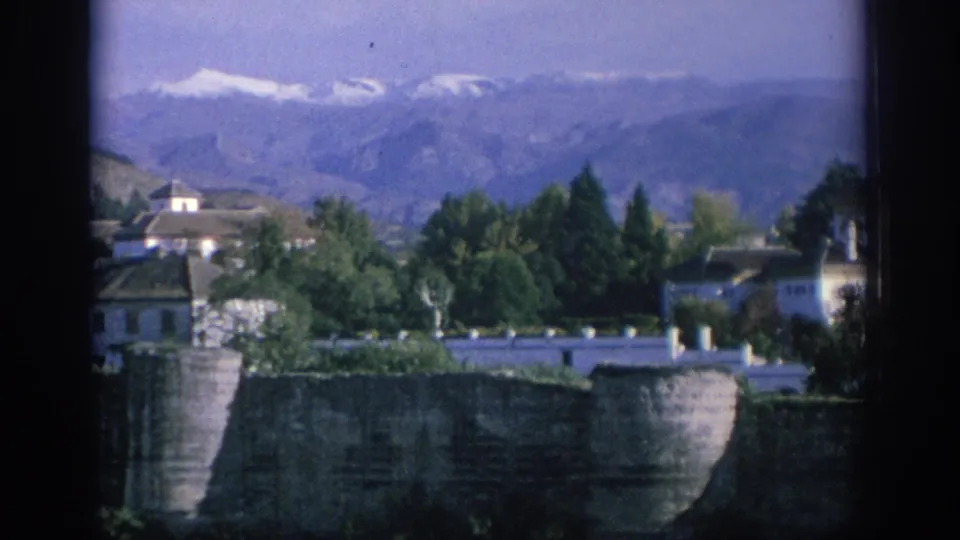 End frame of 1966 Spain 8mm footage showing a quiet exterior landscape with buildings, trees, stonework, and snow-capped mountains.