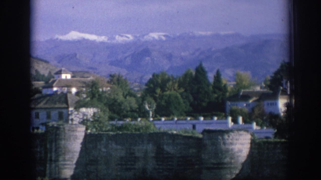 1966 Spain home-movie view across a stone wall toward white buildings, trees, hills, and distant snow-capped mountains.
