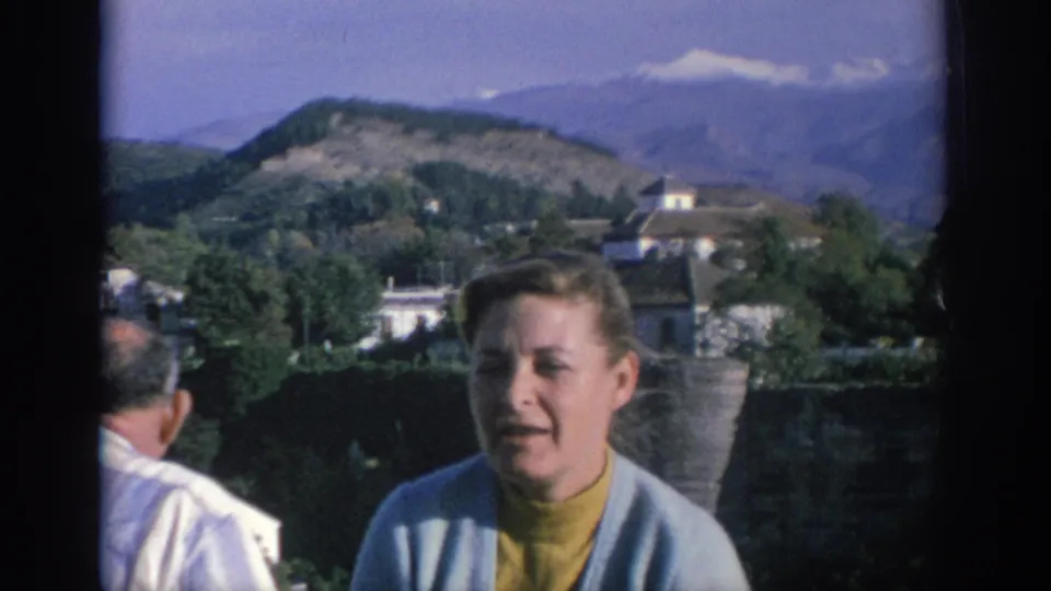 1966 Spain travel footage shows a woman framed close to camera near masonry, with a partial man, trees, buildings, and hills behind her.