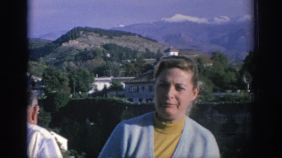 A 1966 Spain home-movie frame shows a woman beside a stone wall, with trees, white buildings, hills, and mountains beyond.