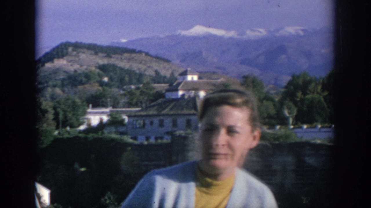 In Spain, 1966, a blond woman in a yellow turtleneck and light cardigan faces the camera with white buildings and mountains behind her.