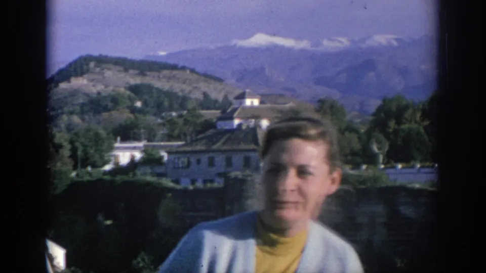 In Spain, 1966, a blond woman in a yellow turtleneck and light cardigan faces the camera with white buildings and mountains behind her.