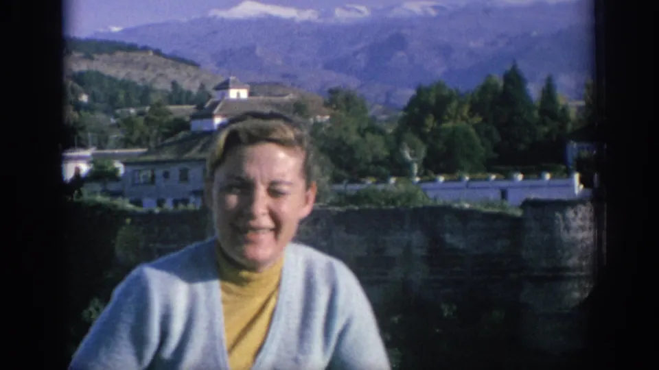 The woman smiles toward the camera in a 1966 Spain travel scene, with whitewashed buildings, trees, rolling hills, and mountains behind her.