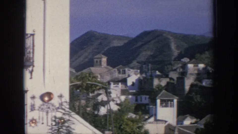 A white exterior wall with a wrought detail and copper cookware display frames a view of rooftops, a tower, and hills in Spain, 1966.