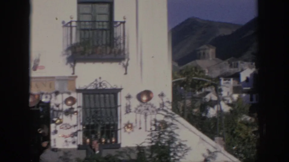 A Spanish town view shows a white facade with wrought-iron balcony, barred window, copperware display, rooftops, and hills beyond.