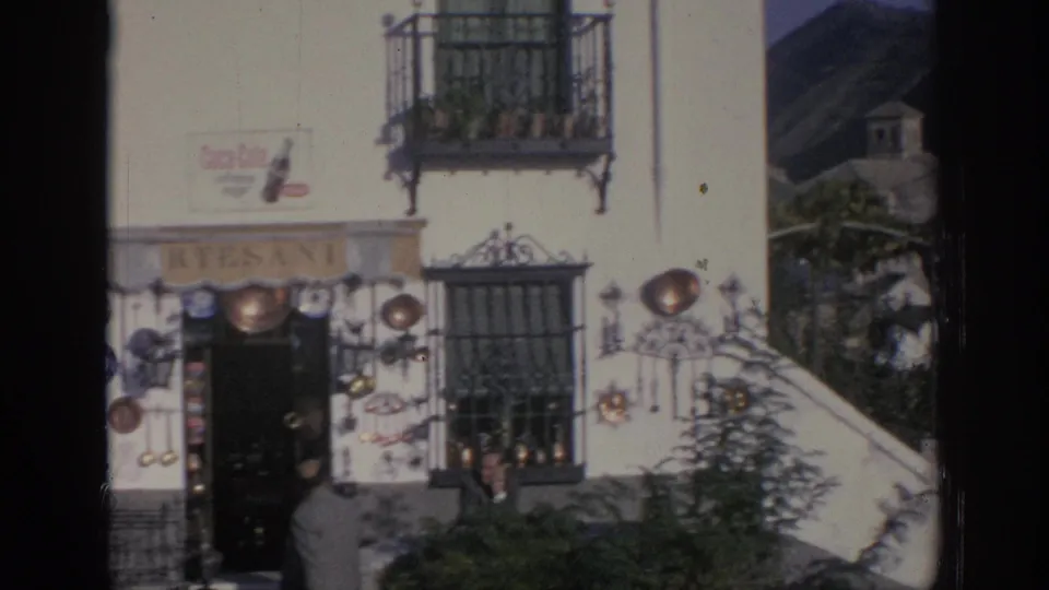 1966 Spain home-movie frame showing a white artisan shop facade with wrought-iron balconies, cookware displays, signage, and people outside.