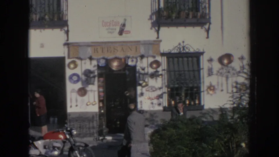 1966 Spain frame of the artisan shop exterior, with displayed cookware, a Coca-Cola sign, a red motorcycle, and several people on the street.