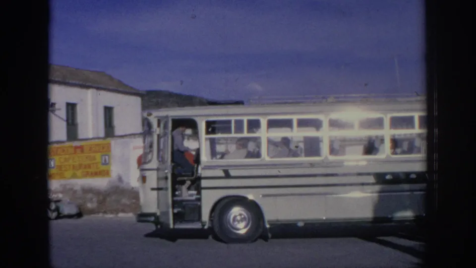 1966 Spain home-movie frame showing a white vintage bus on a street, with passengers visible through side windows and a driver near the open door.