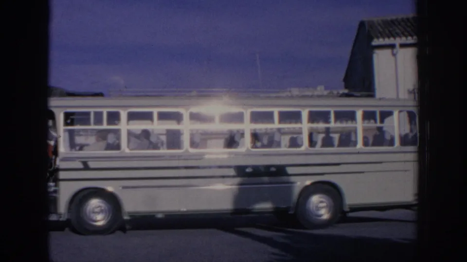 1966 Spain 8mm frame with a side view of a stopped white bus, passengers in the windows, and a dark vertical obstruction in front.