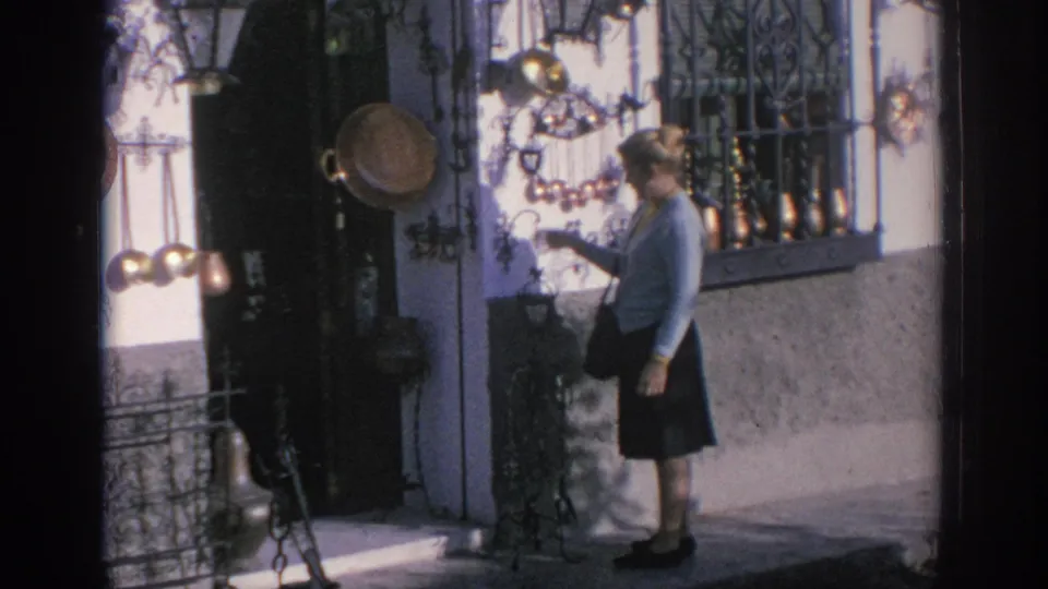 1966 Spain 8mm frame of a woman examining brass or copper items hanging beside a shop doorway on a sunny street.