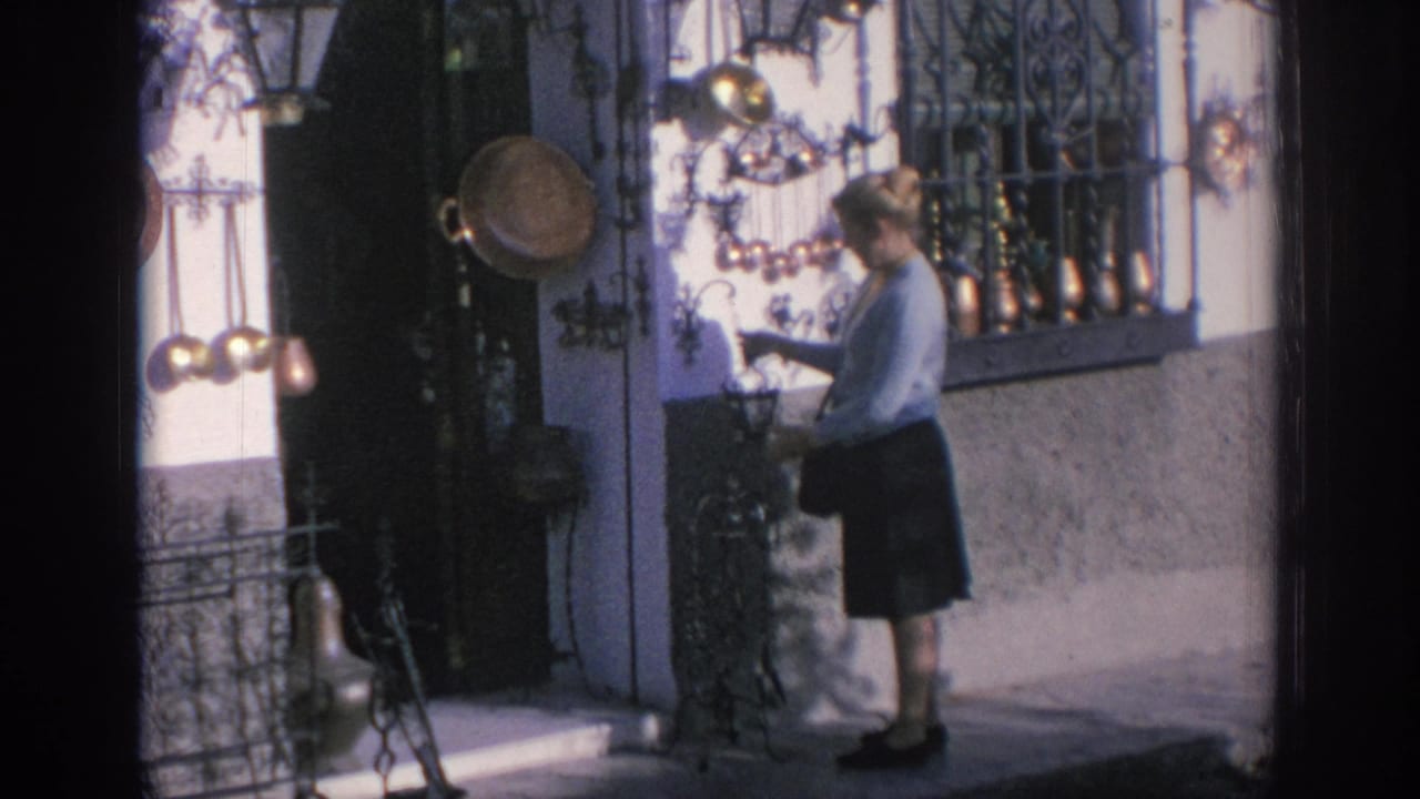 In Spain, 1966, a woman in a light blue sweater browses hanging metal decorations outside a shopfront.