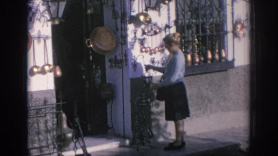 In Spain, 1966, a woman in a light blue sweater browses hanging metal decorations outside a shopfront.