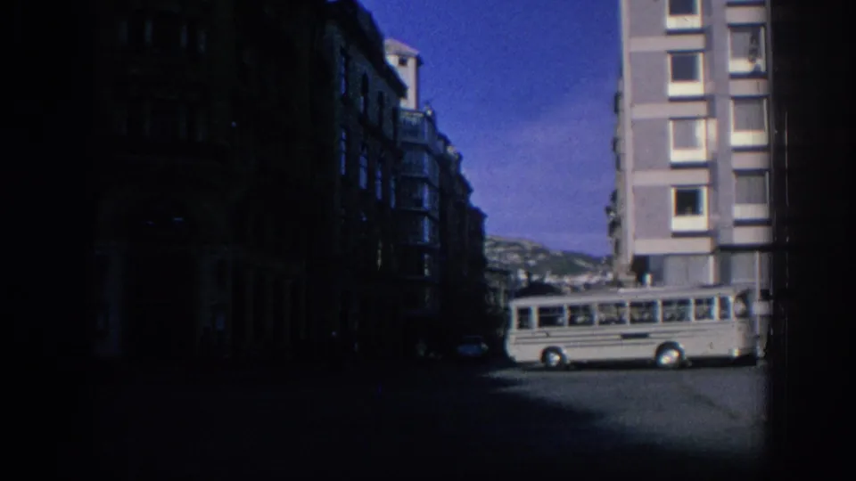 A grey bus passes through a Spanish city street in 1966, with tall buildings and bright sky visible.