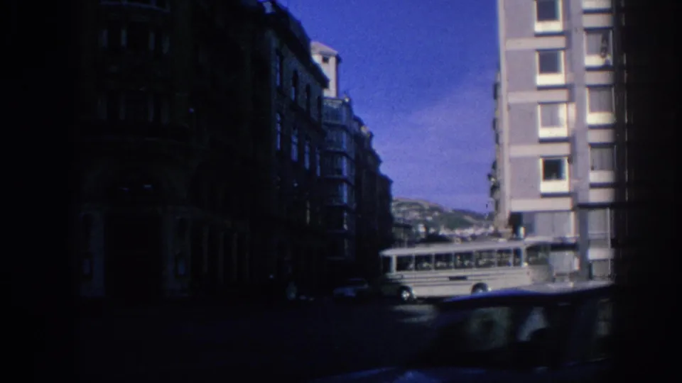 A grey bus moves along a Spanish city street in 1966, framed by dark buildings and a bright sky.