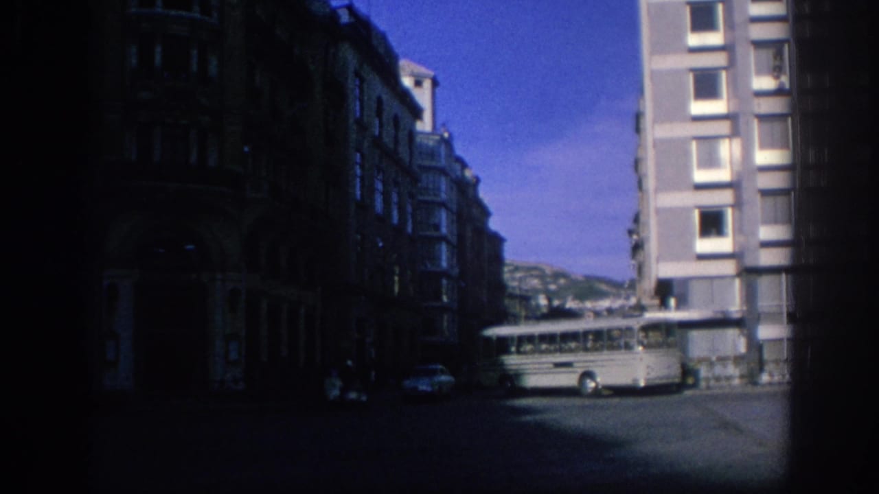1966 Spain home-movie frame showing a narrow urban street, white bus, modern apartment block, and older dark facades.