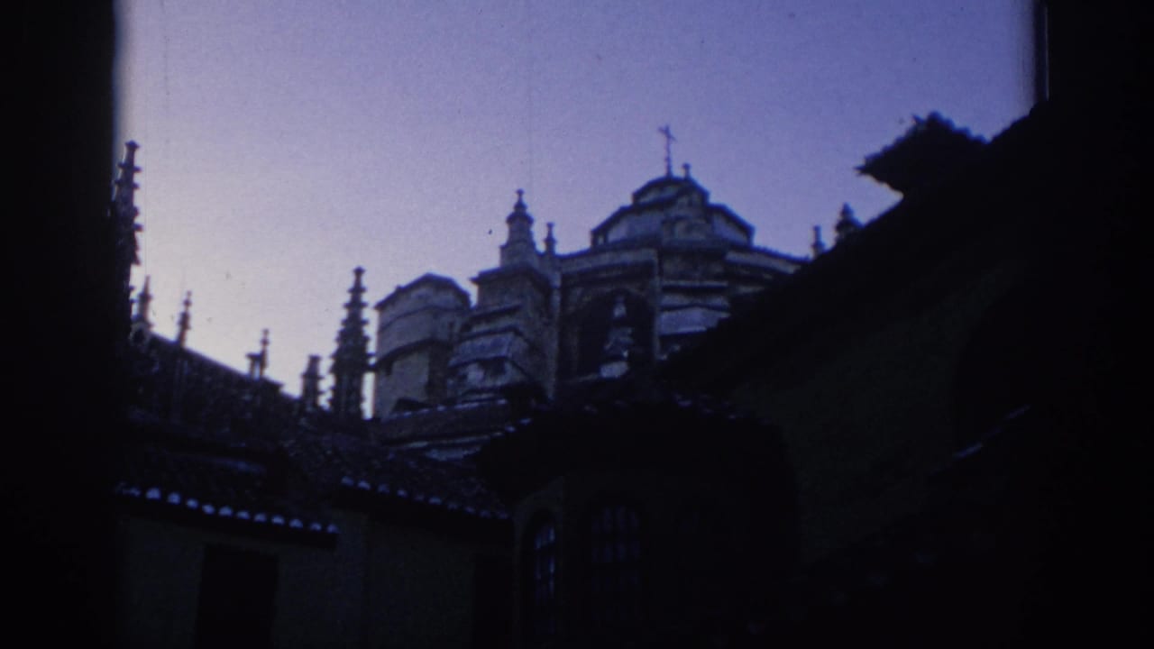 1966 Spain 8mm frame showing dark foreground edges around old rooflines and an ornate building facade, with cool color shift and soft focus.