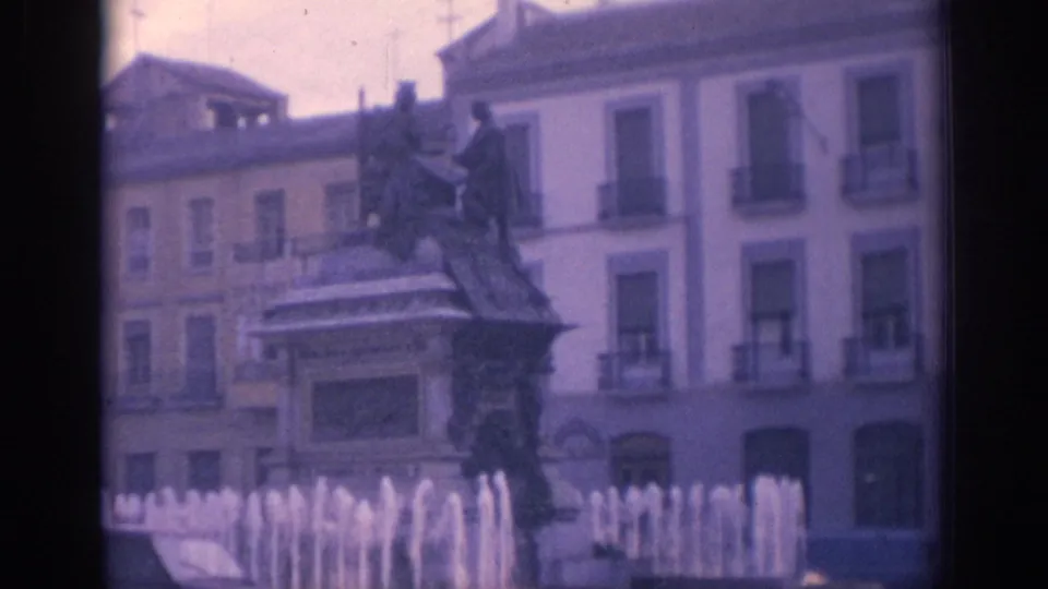 1966 Spain 8mm frame showing the upper statue monument, fountain spray, and apartment-style facades around the square.