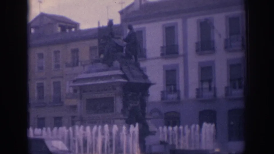 1966 Spain 8mm frame of a public square fountain and statue monument, with balconies and pale building facades behind.