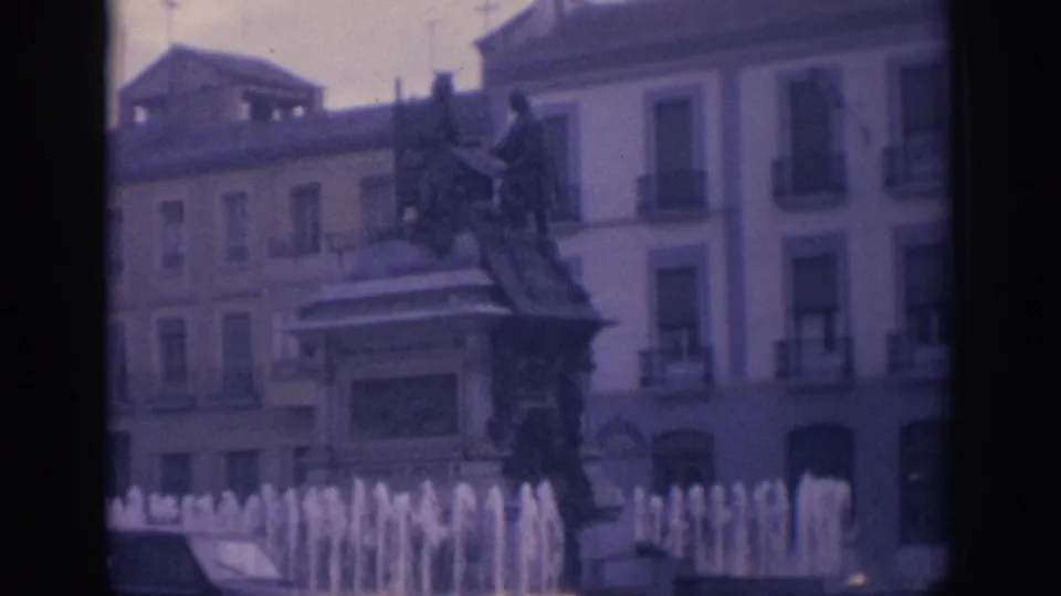 1966 Spain home-movie frame showing a public fountain with a statue before multi-story urban facades, seen in soft 8mm focus.