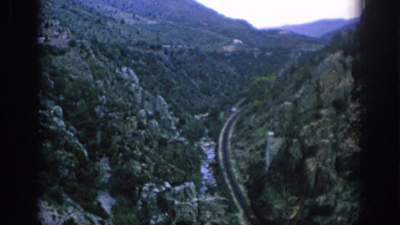 1966 Spain travel footage frame showing a rail line curving through a steep green mountain valley with rocky cliffs and a riverbed below.