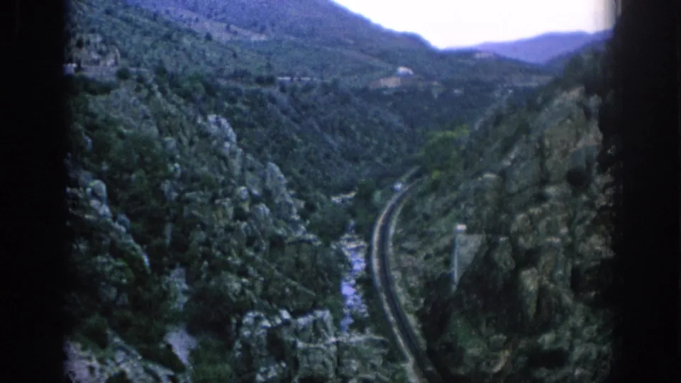 1966 Spain travel footage frame showing a rail line curving through a steep green mountain valley with rocky cliffs and a riverbed below.