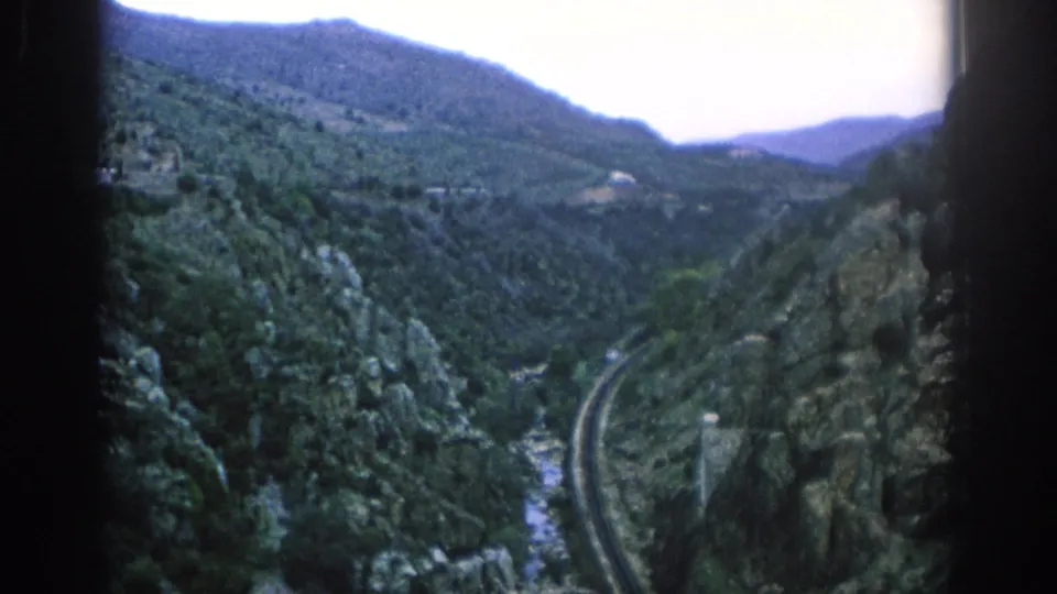1966 Spain 8mm frame with railway tracks following a riverbed between forested rocky slopes and distant mountains.