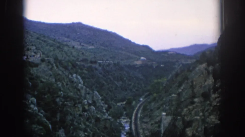 1966 Spain home-movie still showing a curving railway in a rugged mountain valley, framed by dark film borders and soft color.