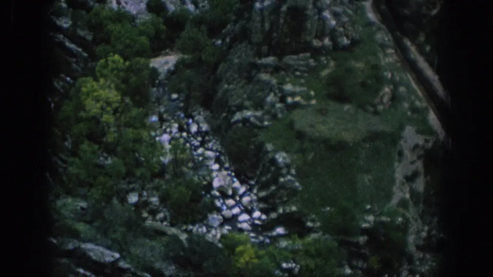 1966 Spain 8mm travel view of a green valley floor with scattered rocks, trees, a stony stream, and a track or road along the side.