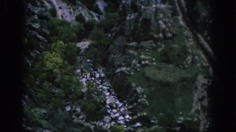 1966 Spain 8mm frame showing a rocky green ravine, bright stream stones, trees, and a railway or road line near the frame edge.