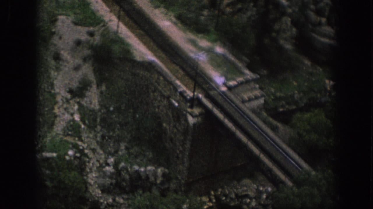 1966 Spain 8mm frame looking down at a weathered stone bridge over a rocky stream in an overgrown rural setting.