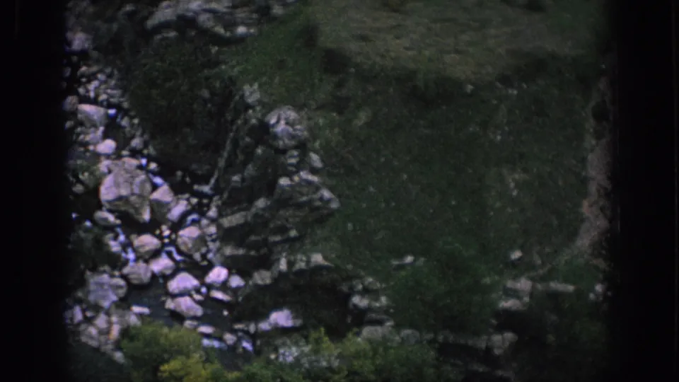 1966 Spain 8mm frame looking down at a rocky stream channel with grass, stones, and overgrown banks near the bridge.