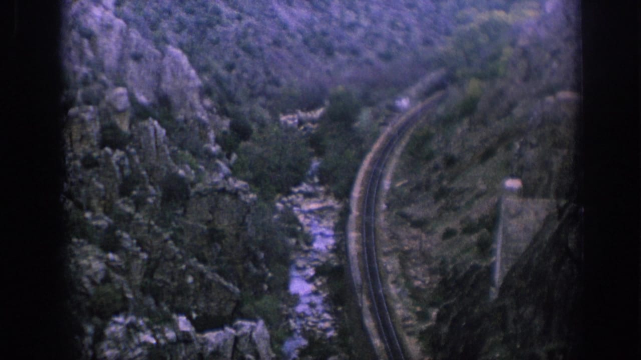 Winding railway track beside a narrow rocky stream in a mountain valley.