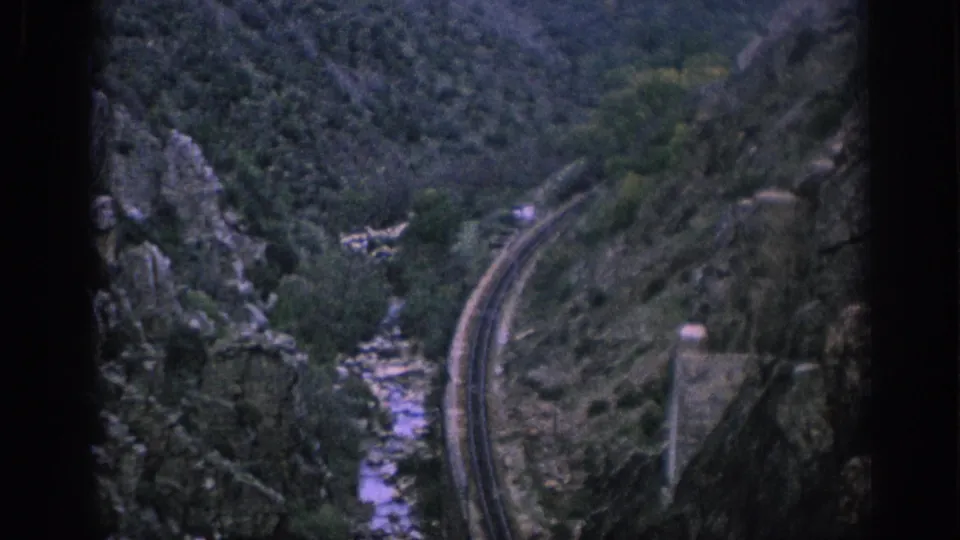 Spain 1966 8mm frame with a railway curve, rocky slopes, and a narrow stream running through the valley floor.