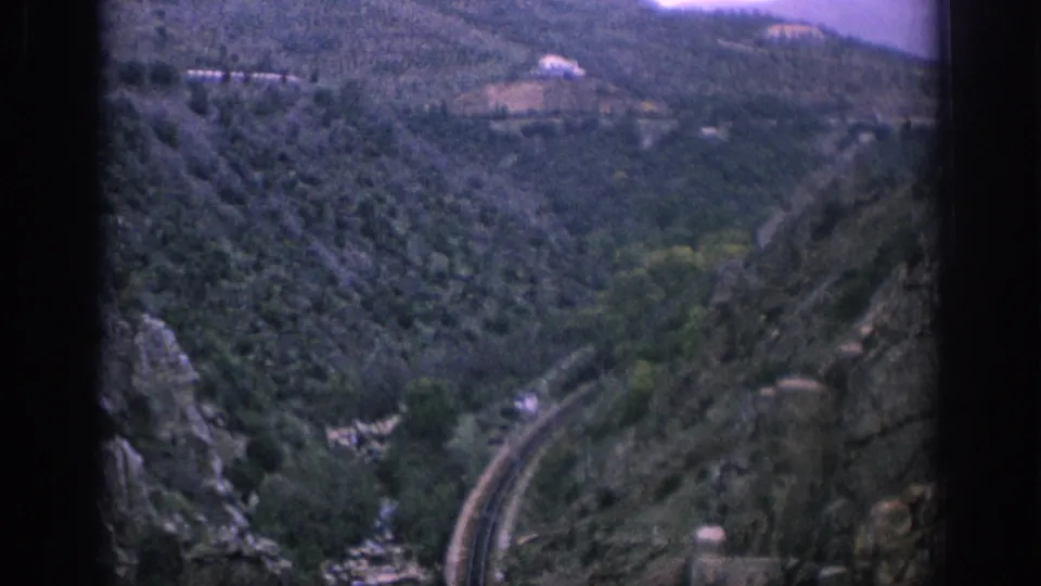 Spain 1966 home-movie frame showing a wider mountain valley, a curving rail line, a stream bed, and small distant hillside structures.