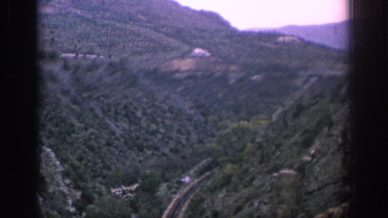 1966 Spain home-movie view of a mountain valley, green slopes, and a curving rail line below a pale sky.