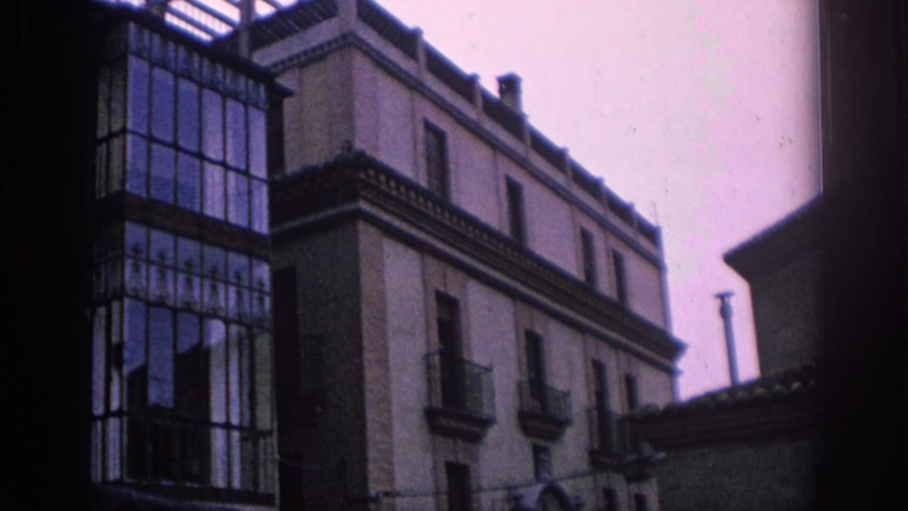 1966 Spain street-level view of a pale building facade beside a tall glass balcony enclosure and tiled roof edges.